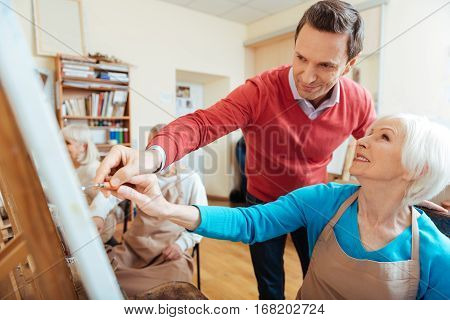 Good advice. Smiling professional artist helping elderly woman while spending time in painting studio and drawing a picture.