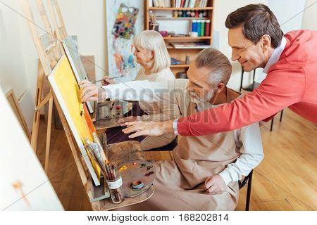 Perfect advice. Overjoyed professional artist helping elderly man while spending time in painting studio and drawing a picture.