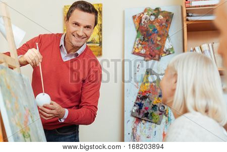 Listen attentively. Smiling handsome young man teaching people and spending time in painting studio while helping them to draw.