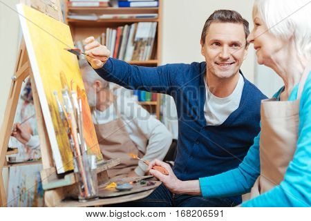 Friendly teacher. Overjoyed handsome young artist helping elderly woman while he painting and spending time in painting studio