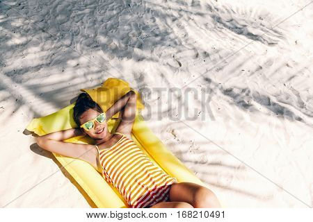 Child girl in yellow swimwear and sunglasses chilling on lilo under palm tree on sand at the beach. Tropical island. Summer holiday idyllic. Top view.