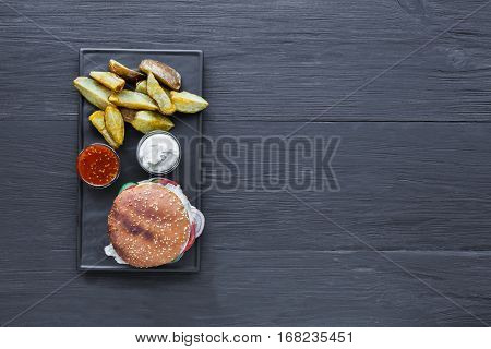 Fast food restaurant dish top view with copy space. Meat cheese burger, potato chips and wedges. Take away set on dark black slate plate on wood background. Hamburger and spicy tomato sauce.