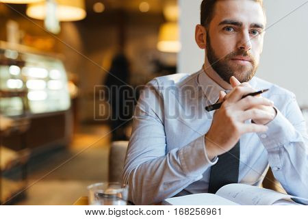Portrait of young bearded businessman in shirt and tie sitting at cafe table with arms clasped under his chin and looking at camera pensively.