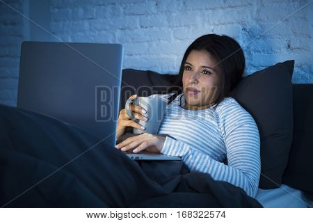 young beautiful hispanic woman in pajamas on bed at home bedroom working happy with laptop computer late at night or using the internet chat and social network in technology addiction concept
