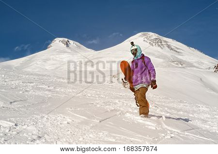 Stylishly dressed snowboarder stands and walks on mountain slopes of an extinct volcano Elbrus on a background of pure blue sky