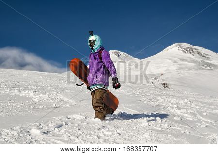 Stylishly dressed snowboarder stands and walks on mountain slopes of an extinct volcano Elbrus on a background of pure blue sky