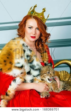 Red-haired woman in red dress, cloak and with crown on head sits on couch holding lynx cub.