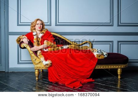 Blonde woman in red dress and cloak lies on couch in room.