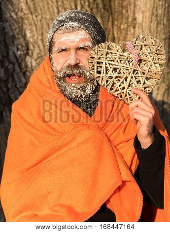 Frozen smiling man with squinty eyes bearded hipster with beard and moustache covered with white frost wrapped in orange blanket with wicker heart on winter day outdoors on natural background