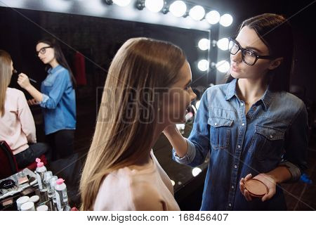 Natural look. Nice positive female makeup artist holding face powder and applying it on the cheek of her client with a makeup brush while standing in front of her