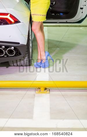 Legs in blue high-heel shoes of woman in yellow dress standing near modern white car.