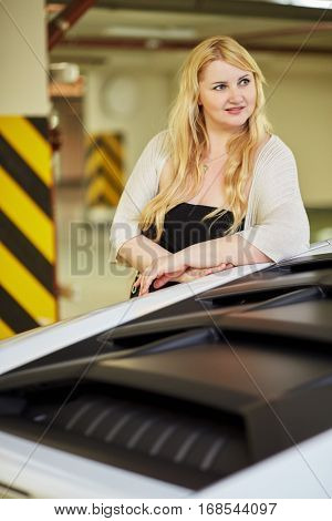 Young blonde woman stands leaning on foof of modern white car at underground parking.
