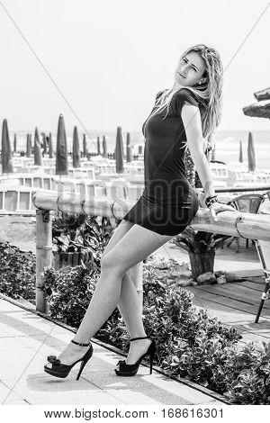 Feminine seduction. Blonde model girl outdoors. Black and white. A beautiful blond girl is leaning against a fence. She wears a black dress and shoes with heels. Black and white.