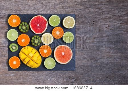 Raw fruit rich in vitamin C on the black slate platter: oranges grapefruits lemons limes kiwis slices, top view, copy space for text