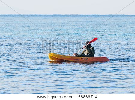 Fisherman In Green Jumpsuit Floats In Orange Kayak On Sea