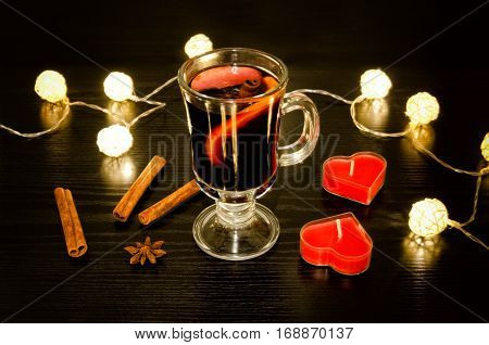 Mug of mulled wine with spices candles in the shape of a heart cinnamon sticks star anise. Illumination of rattan lanterns on a black wooden table