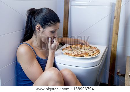 young sad and depressed bulimic woman feeling sick guilty sitting at the floor of the toilet leaning on WC eating pizza in nutrition disorder bulimia concept