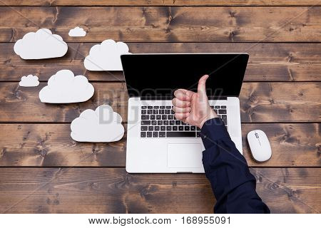 Cloud computing technology concept with white fluffy clouds next to the laptop. Man with thimbs up over the keyboard uploading data on a wooden table.