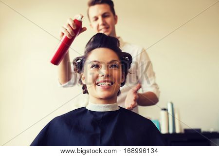 beauty, hairstyle and people concept - happy young woman with hairdresser with hair spray fixating hairdo at salon
