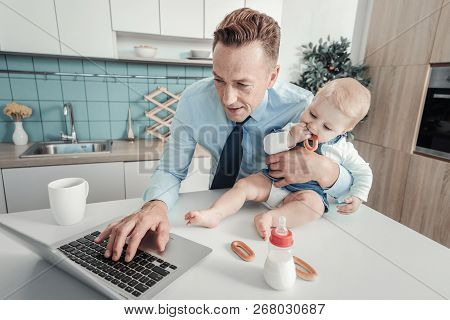 Pleasant Busy Man Hugging A Nursling And Using The Laptop.