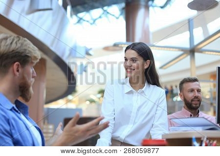 Group Of Young People In Casual Wear Sitting At The Office Desk And Discussing Something While Looki