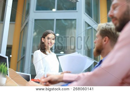 Group Of Young People In Casual Wear Sitting At The Office Desk And Discussing Something While Looki
