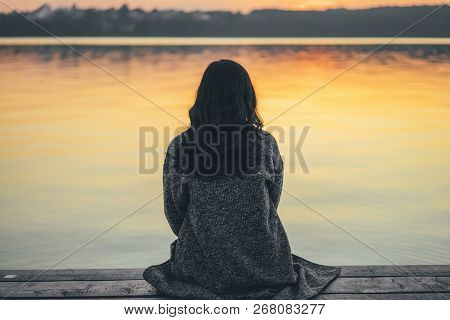 Back View Of Long Hair Brunette Girl Sitting On The Pier Near Lake At Sunset Time.