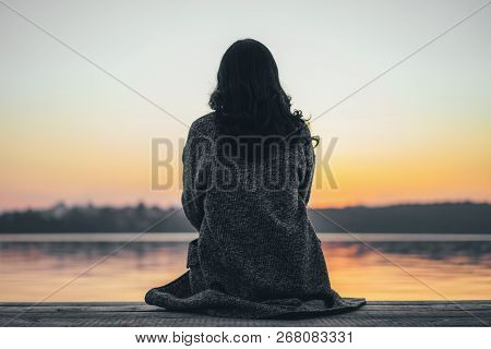 Back View Of Long Hair Brunette Girl Sitting On The Pier Near Lake At Sunset Time.