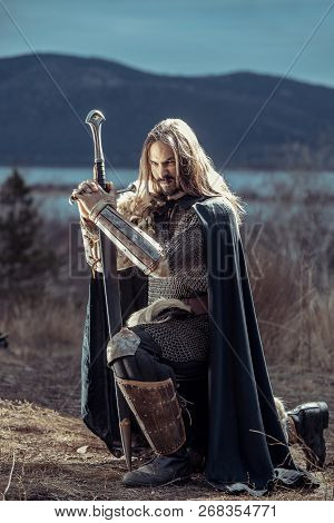 Long Haired Knight With The Two-handed Sword. Medieval Rural Background.