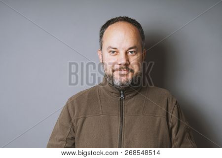 Middle Aged Man In His 40s With Short Dark Hair And Graying Beard Smiling Against Gray Wall With Cop