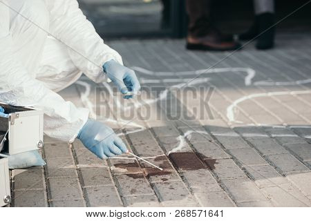 Male Criminologist In Protective Suit And Latex Gloves Collecting Blood Sample At Crime Scene
