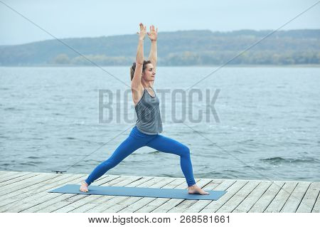 Beautiful Young Woman Practices Yoga Asana Virabhadrasana 1 - Warrior Pose 1 On The Wooden Deck Near