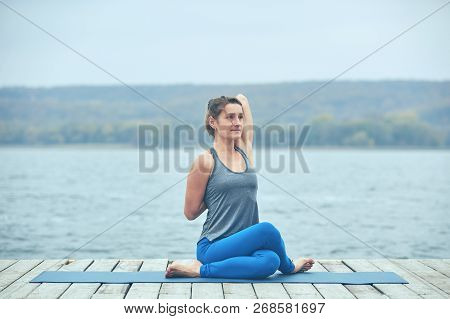 Beautiful Young Woman Practices Yoga Asana Gomukhasana - Cow Face Pose On The Wooden Deck Near The L