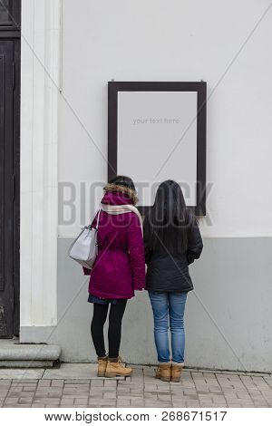 Two Women Stand At The Poster And Read The Text. View From The Back. Advertising Concept.
