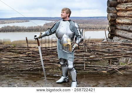 Knight In Armor On The Background Of A Wicker Fence, A Wooden House And The Expanses Of The Motherla