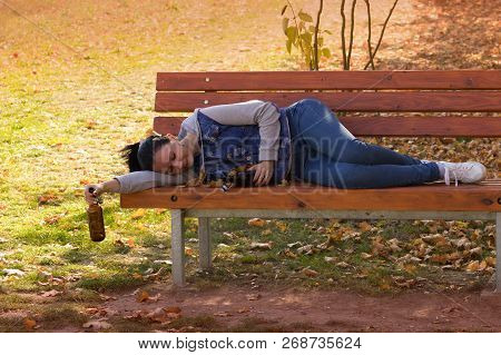 A Drunk Young Woman Sleeping On A Bench In The Day.a Young Woman Holding A Bottle Of Alcohol In Her 