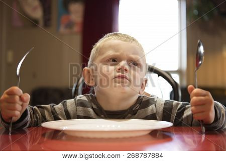 A Upset Little Boy Waiting For Dinner While Holding A Fork And A Spoon