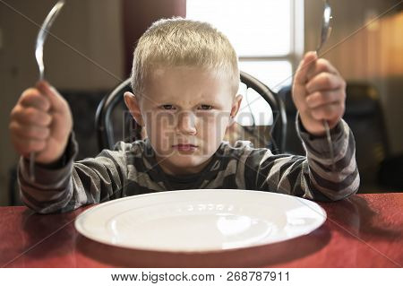 A Upset Little Boy Waiting For Dinner While Holding A Fork And A Spoon