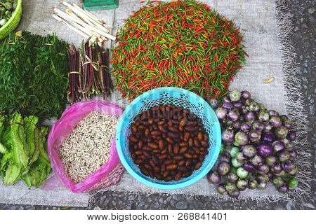 Hot Red And Green Peppers, Eggplants, Beans, Greens, And Edible Larva And Insect Pupa At A Market In