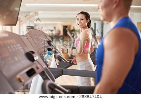 Pretty Smiling Asian Woman Looking At Handsome Man On Threadmill Next To Her