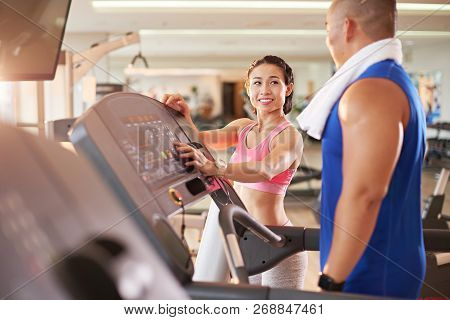 Pretty Female Fitness Trainer Explaining Interface Of Threadmill To Her New Client