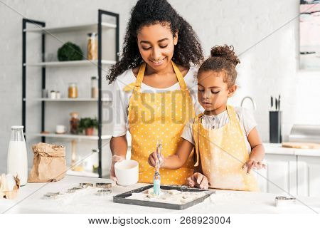 Smiling African American Mother And Daughter Applying Oil On Unbaked Cookies In Kitchen
