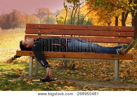 A Drunk Young Man Sleeping On A Bench In The Day.a Young Man Holding A Bottle Of Alcohol In Her Hand