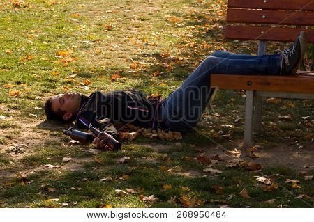 A Drunk Young Man Sleeping On Grass Under The Bench In The Day.a Young Man Holding A Bottle Of Alcoh