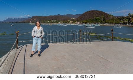 Smiling Tourist With Casual Clothes Posing On The Boardwalk With The Lake With Motorboats, The Pier 
