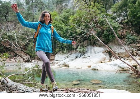 A Woman With A Backpack Is Walking Along The Trunk Of A Fallen Tree. A Girl Crosses A River On A Log