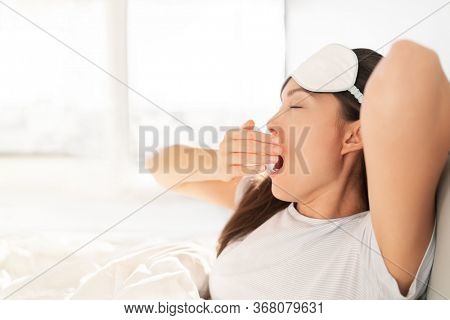 Waking up early morning at sunrise happy woman yawning stretching in bed. Sleeping at home woman waking up in morning sunlight stretching happy after a good night sleep feeling rested wearing eye mask