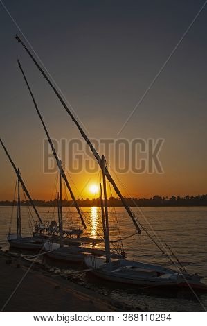 Traditional Egyptian Felluca River Boats Moored On The Nile River Bank With Sunset And Reflection In