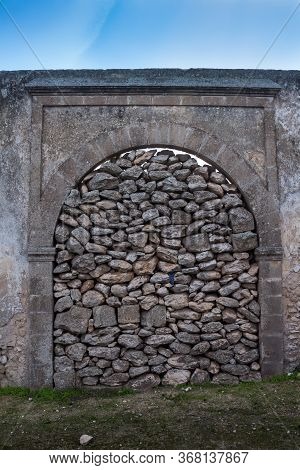 Fence With A Gate With An Arch, Barred With Stones. Part Of An Abandoned Building. Blue Sky With Whi