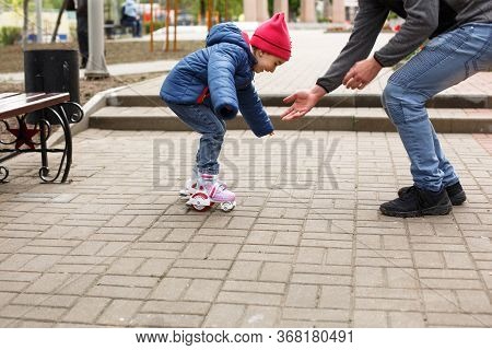 Dad Teaches His Little Daughter To Ride Four-wheeled Roller Skates. The Father Supports The Child By